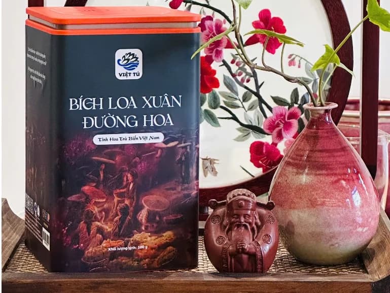 Hộp thiếc trà đặt trên kệ gỗ, cạnh bình hoa gốm và một tượng gốm nhỏ